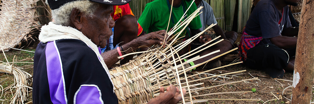 An elderly lady sits weaving a basket by hand using natural leaves
