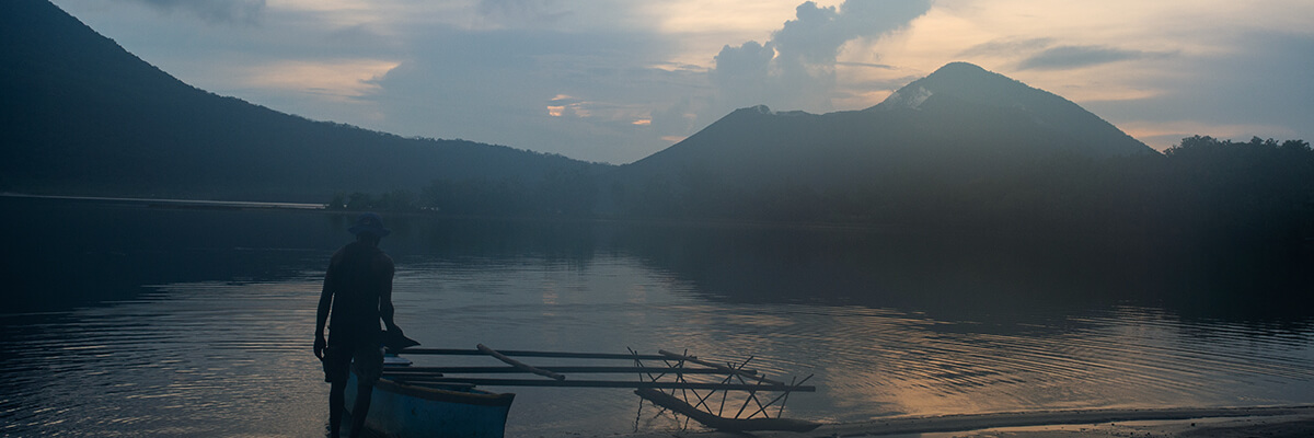 A man tugs his catamaran behind him, towards a misty lake.