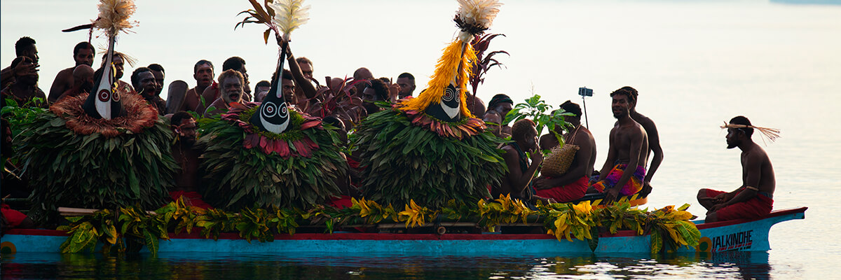 A group of tribespeople sit in a boat, some dressed in traditional clothing