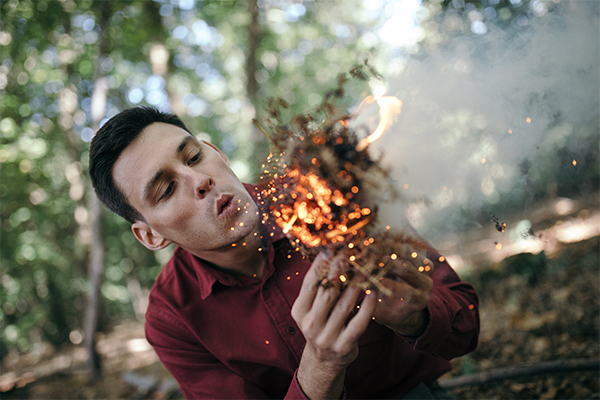 Daniel Hume blows into a bundle of tinder, creating orange glowing embers and  light smoke.