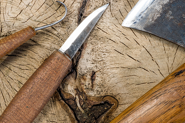A knife, axe and spoon gouge tools lay upon a wooden tree stump.