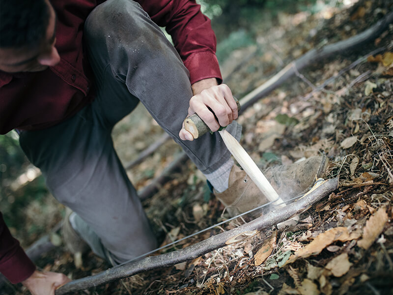 Daniel Hume crouches on one knee, holding a bow in one hand and a wooden drill in the other, using them in tandem to create smoke.