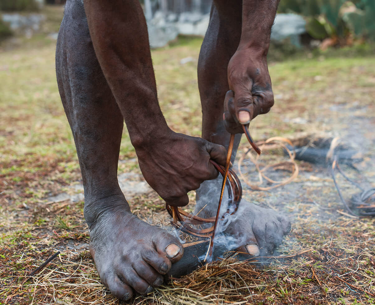 A native tribesman holds a chunk of wood between his feet, and grasps a length of twine in his hands. Rubbing them together, he creates smoke.