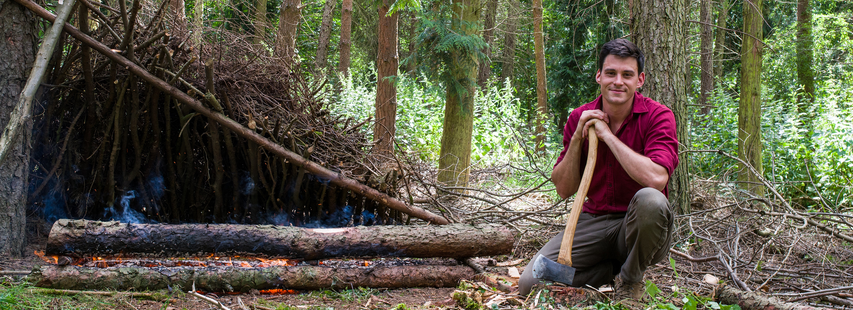Daniel Hume crouches beside a shelter made of sticks, with a long-log fire infront. He rests his hands on an upended axe.